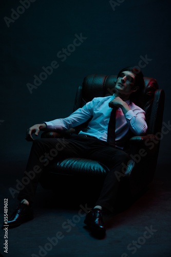 Portrait of a man in blue and red contrasting lighting in a white shirt with a tie and a facial injury, posing sitting in a leather chair. Futuristic neon lighting