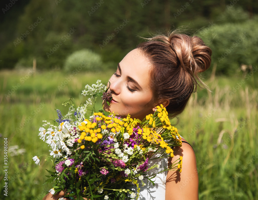 Naklejka premium Summer lifestyle portrait of beautiful young woman smiling and holding bouquet of wild flowers. Standing in the field of grass. Enjoying sun. Happiness and love concept. Сlosed eyes 