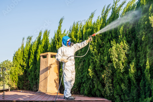 Fumigator applying plant protection products and herbicides to the plants of a house with a garden raised on the roof of a building. The worker is wearing a protective suit against toxic products and 