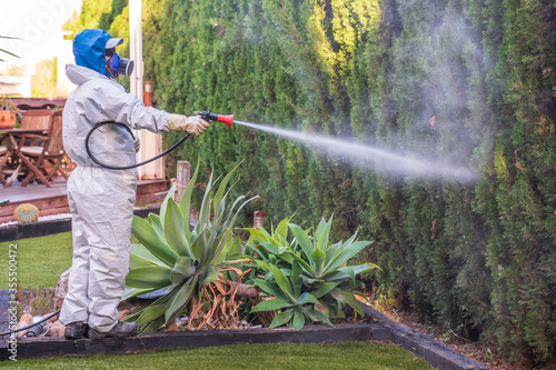 Fumigator applying plant protection products and herbicides to the plants of a garden house next to a Japanese-style stone fountain. The sprayer is wearing a protective mask and a white protective sui photo