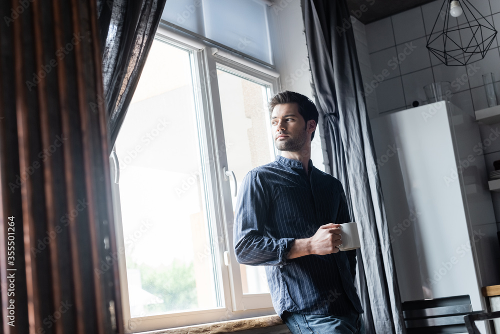 handsome pensive man holding cup of coffee and standing near window at home on self isolation handsome pensive man holding cup of coffee and standing near window at home on self isolation