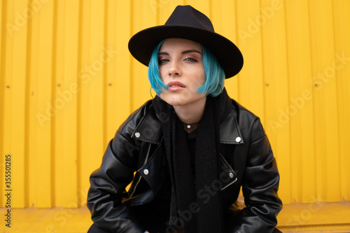 Young woman with blue hair in a black leather jacket and hat sits near an orange wall photo