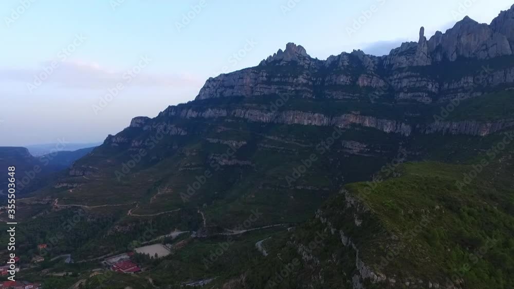 Aerial panning shot of rocky mountains by city in valley against sky, drone flying over landscape during sunset - Montserrat, Spain Aerial panning shot of rocky mountains by city in valley against sky, drone flying over landscape during sunset - Montserrat, Spain