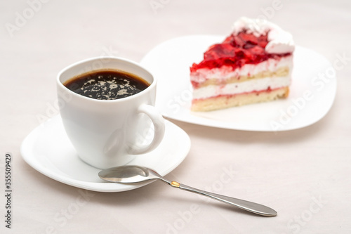 A cup of black coffee on a saucer with a spoon on a table. On a background a piece of cake on a plate