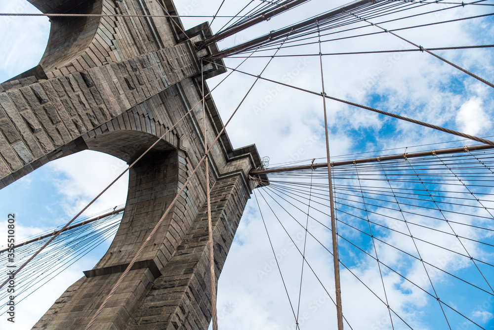 Fototapeta premium Ikoniczny Brooklyn Bridge w Nowym Jorku, USA. Kamienna architektura na dużą skalę z flagą kraju na częściowo zachmurzonym niebie.