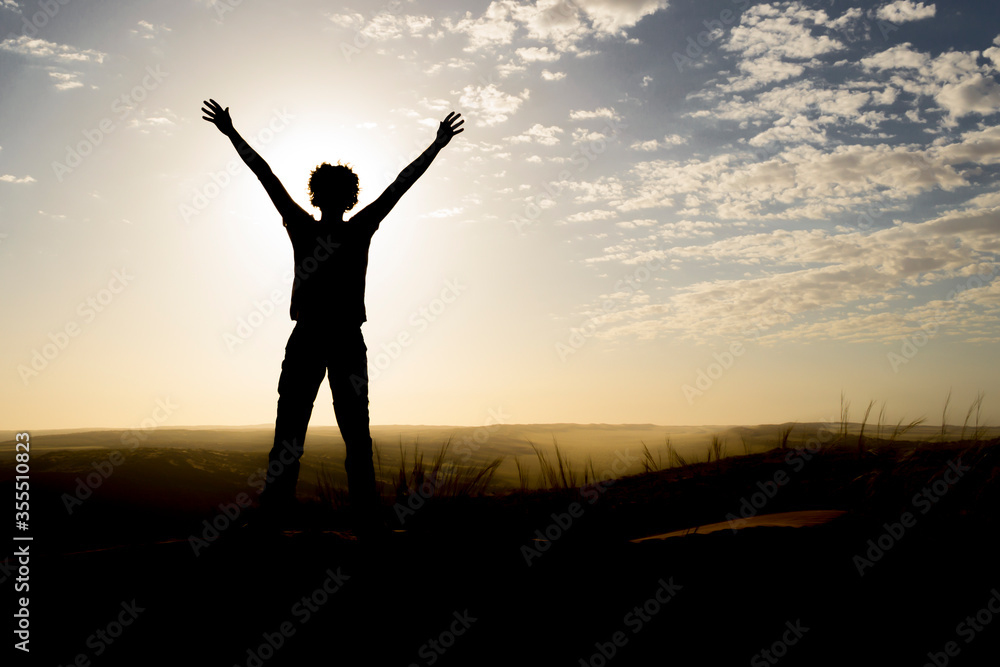Silhouette of person standing on a dune during sunrise, arms in the air.