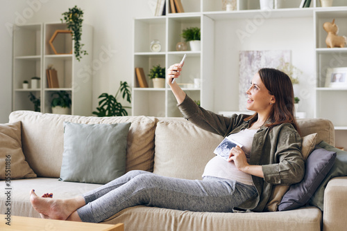 Happy pregnant woman with ultrasonic picture of her baby making selfie on couch