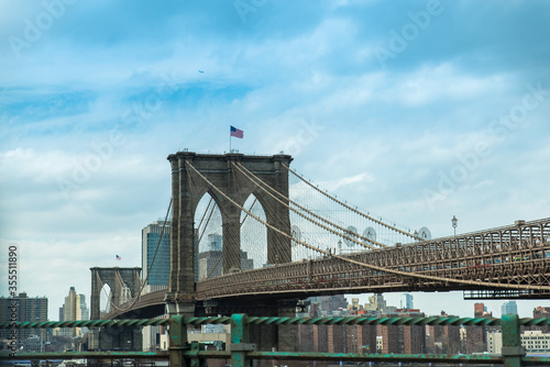 Fototapeta Naklejka Na Ścianę i Meble -  Iconico puente de Brooklyn en Nueva York, Usa. Arquitectura a gran escala de piedra con bandera ddel pais sobre cielo parcialmente nubado azul. 