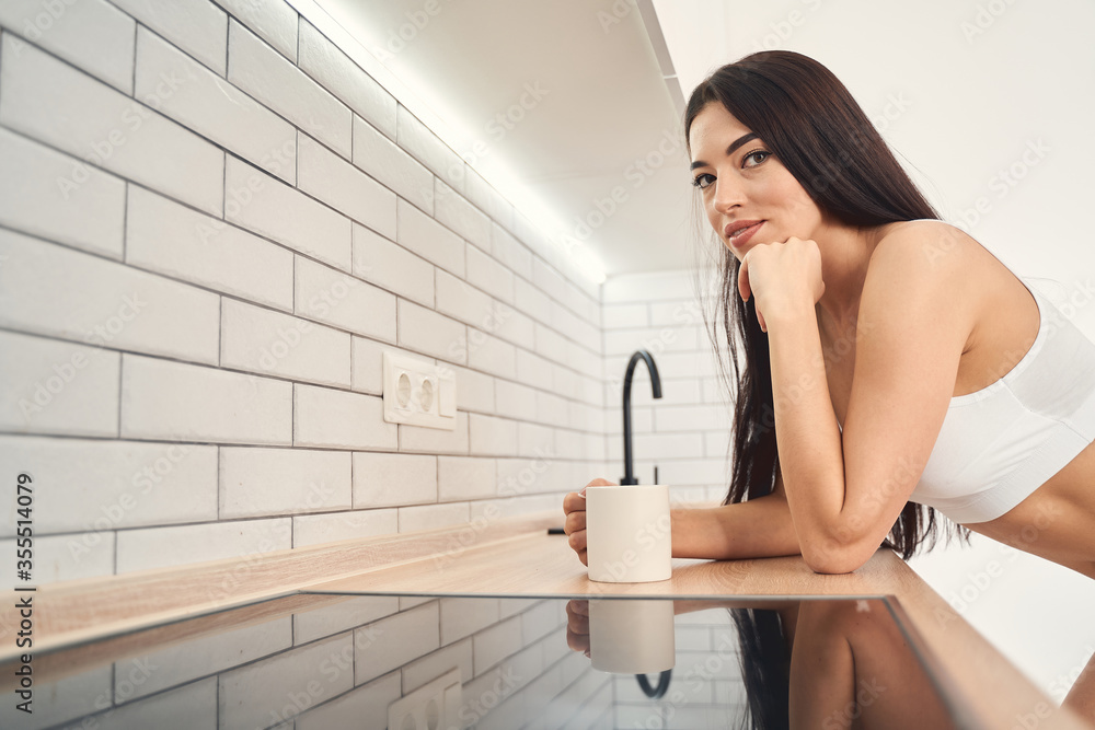 Smiling young woman bending over the kitchen bench holding cup of beverage Smiling young woman bending over the kitchen bench holding cup of beverage