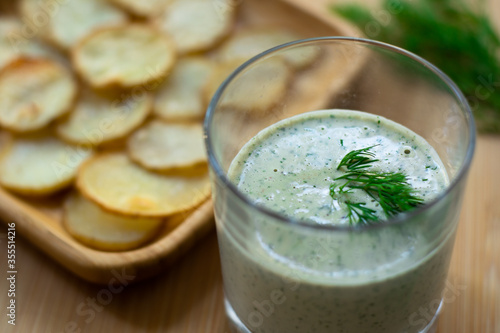 Dill and sour cream dip for homemade potato chips made vegan: healthy snack with dill, tahini, water, vinegar and lemon juice in a glass. Thinly sliced and baked potatoes in the back