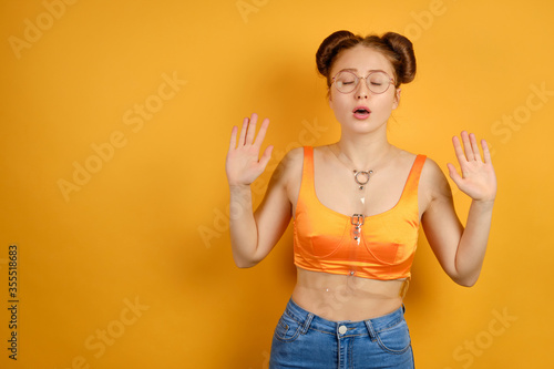 A red-haired girl in an orange top and glasses stands on a yellow background, closing eyes and raising her hands, palms forward
