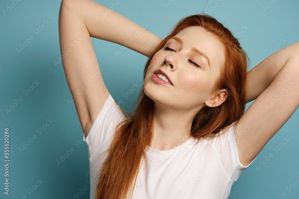 A red-haired girl in a white T-shirt stands on a blue background, closing her eyes and throwing her hands behind her head A red-haired girl in a white T-shirt stands on a blue background, closing her eyes and throwing her hands behind her head