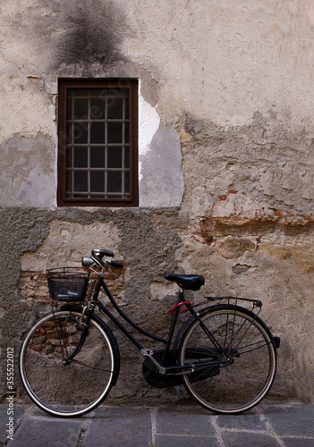 An old bicycle with metal basket rests against a weathered cement wall in Lucca, Italy © Claudia