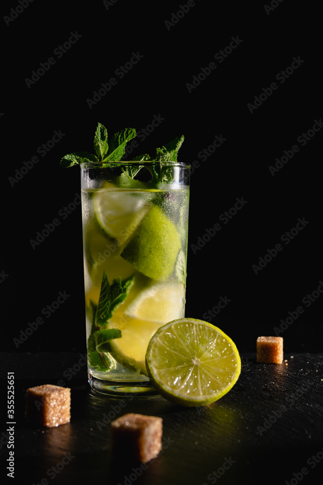 Fototapeta premium A glass of cold mojito cocktail with mint leaves and cubes of cane sugar on a black background.