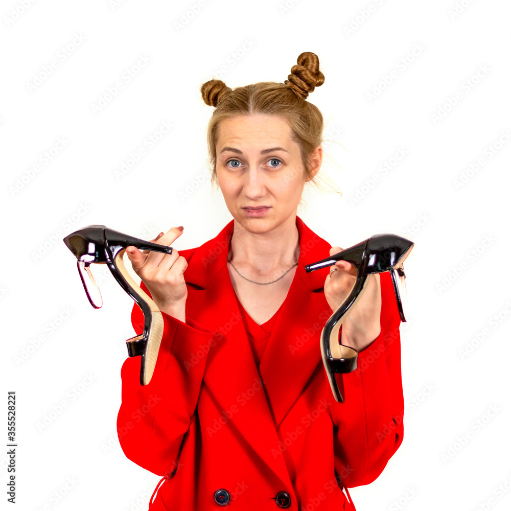 A beautiful young stylish girl looks disappointed and sad at the camera dressed in a red coat and holding women's summer sandals in her hands. The concept of women's happiness, clothing, emotion of A beautiful young stylish girl looks disappointed and sad at the camera dressed in a red coat and holding women's summer sandals in her hands. The concept of women's happiness, clothing, emotion of