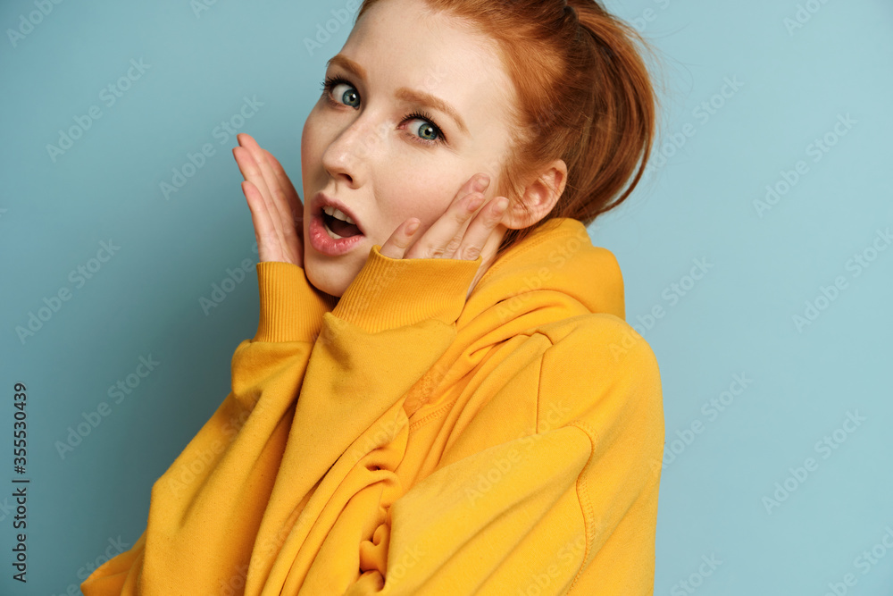 A red-haired girl in a yellow sweatshirt stands on a blue background and looks in surprise at the camera, raising palms to face A red-haired girl in a yellow sweatshirt stands on a blue background and looks in surprise at the camera, raising palms to face