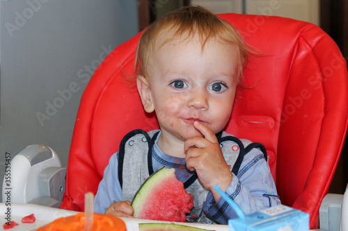 The baby is sitting in a red child's chair and eating a watermelon. He's all dirty, but he's eating himself. photo