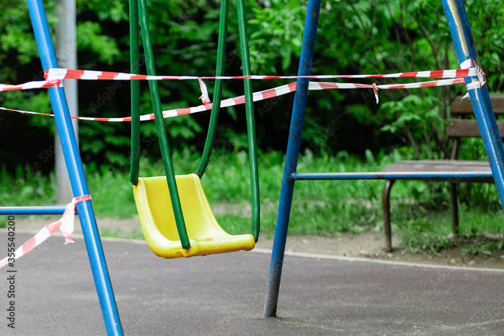 Fototapeta premium Yellow children's swing on a sports playground wrapped with red barrier tape. Prohibition of outdoor walks, prevention of the coronavirus influenza virus covid-19. infection. selective focus