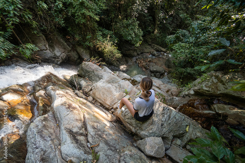 Beautiful young slim blonde woman is sitting on a rock and enjoy of tropical waterfall in jungle. Travel concept.