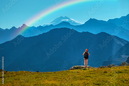 Girl traveler with backpack stands on a stone back to the viewer. She looks at the snow-capped top. Picturesque cloudless landscape with a rainbow. Wallpaper for concept of business thinking. extreme