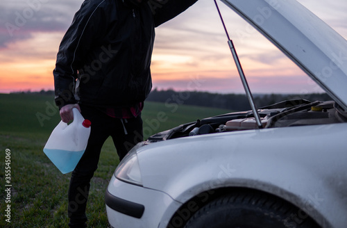 Man standing near car with open hood and holding a bottle with blue fluid on sunset. Car breakdown. Confused man stands with open hood with selective soft focus. © Volodymyr