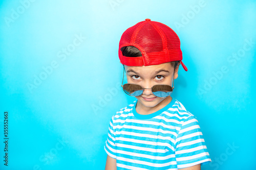 Crazy boy wearing a striped t-shirt and sunglasses with a baseball cap on his head over an isolated blue studio background. Emotion, expression. Copy space for text.