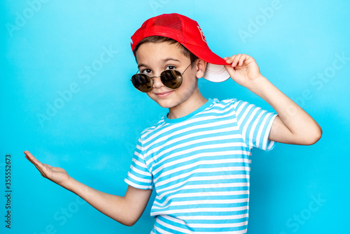 Crazy boy wearing a striped t-shirt and sunglasses with a baseball cap on his head over an isolated blue studio background. Emotion, expression. Copy space for text.
