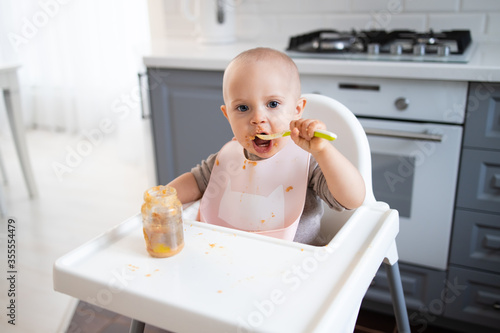 baby sits in a feeding chairgirl in the kitchen and eats photo