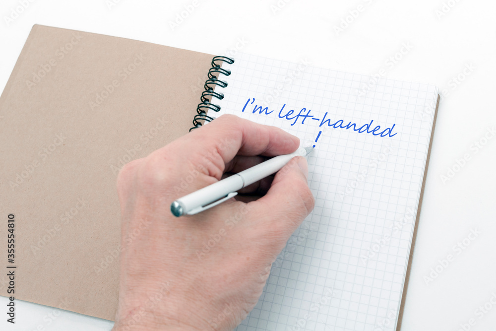 Left-handed day concept. Above the open notebook, the left hand with a pen. The inscription in the notebook : Left-handed day concept. Above the open notebook, the left hand with a pen. The inscription in the notebook :