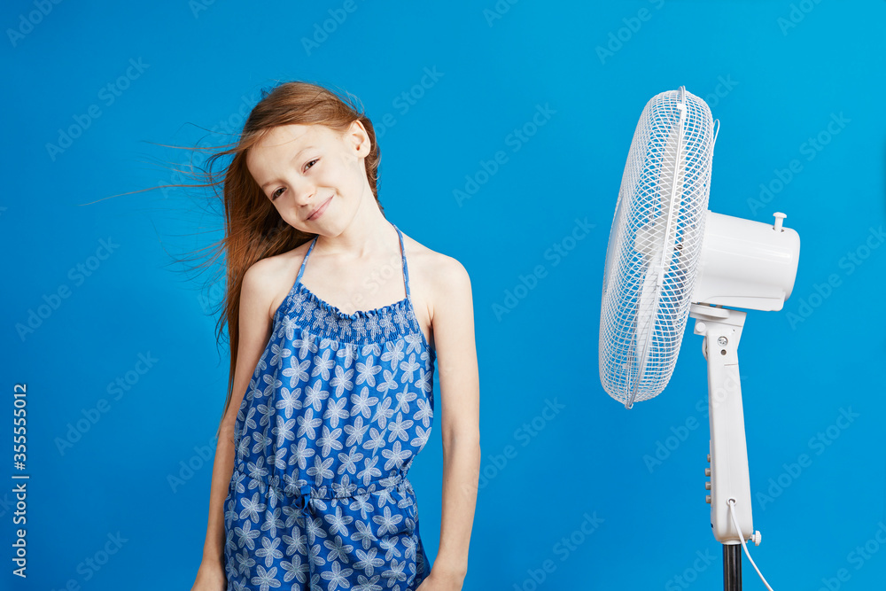 jolie jeune enfant fille caucasienne aux cheveux longs châtains devant un ventilateur sur fond bleu de studio jolie jeune enfant fille caucasienne aux cheveux longs châtains devant un ventilateur sur fond bleu de studio