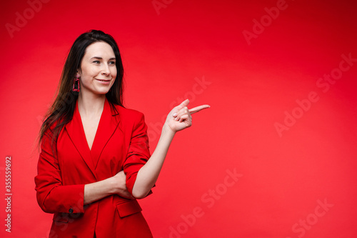 Stock photo portrait of a cheerful smiling brunette in red jacket pointing with index finger at blank space on plain red background. Adult woman with long dark hair in red showing something in the air