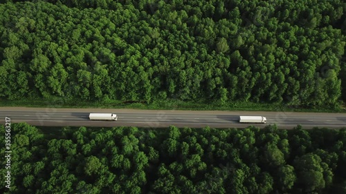 Two semi trucks with white trailer driving / traveling on the asphalt straight road at dense flat forest, highway top view follow vehicle aerial footage at summer sunset / Freeway truck traffic