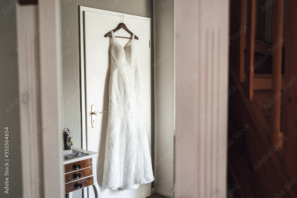 White wedding gown hanging on hanger on white doors. Bride's wedding dress in her room. Wedding day concept. White wedding gown hanging on hanger on white doors. Bride's wedding dress in her room. Wedding day concept.