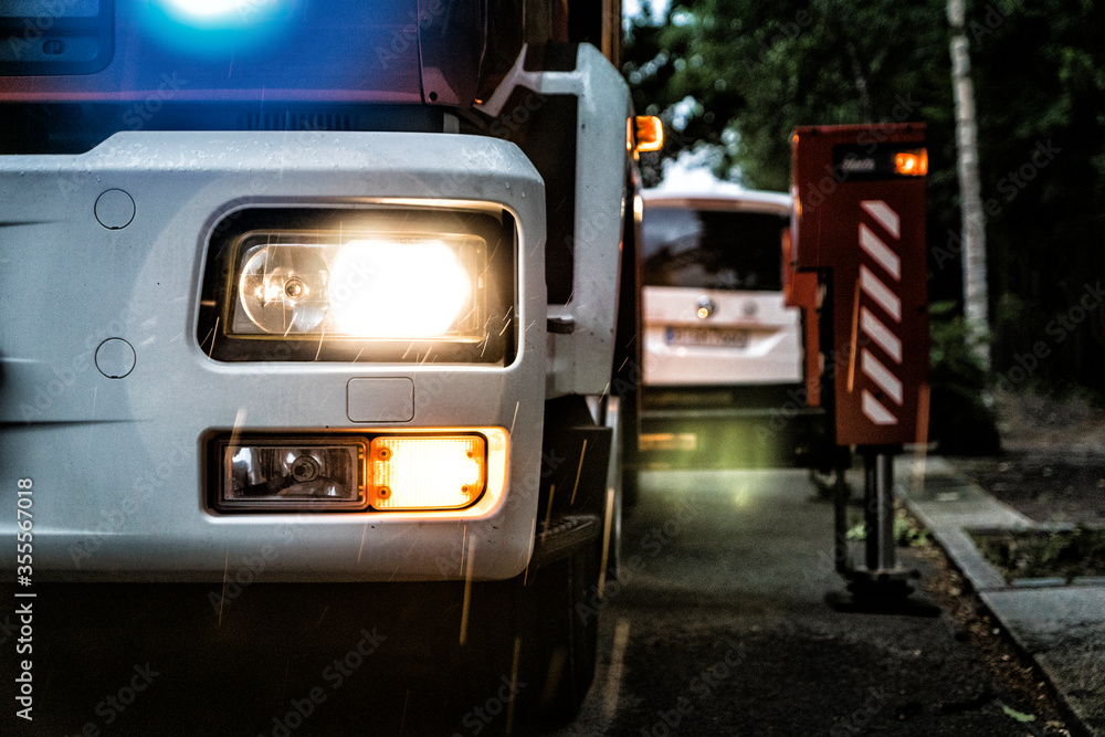 Frontbeleuchtung eines Feuerwehrautos während eines Einsatzes bei Dämmerung mit Blaulicht, Warnblinklicht und Scheinwerfern. Im Hintergrund sind die ausgefahrenen Stützen der Drehleiter erkennbar.