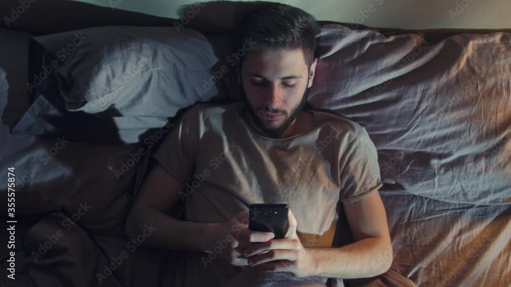 Sleepless night. Social media addiction. Bored man reading feed on phone in bed. Sleepless night. Social media addiction. Bored man reading feed on phone in bed.