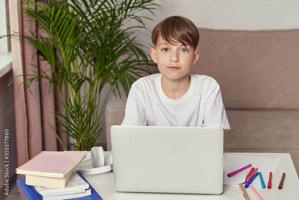 Beautiful boy in white clothes. Looks at the camera. On the table is a laptop Beautiful boy in white clothes. Looks at the camera. On the table is a laptop