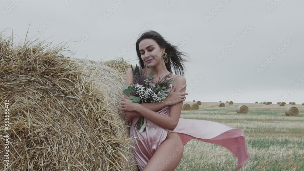 Beautiful naked woman in dress with flowers poses at haystack in windy field Beautiful naked woman in dress with flowers poses at haystack in windy field