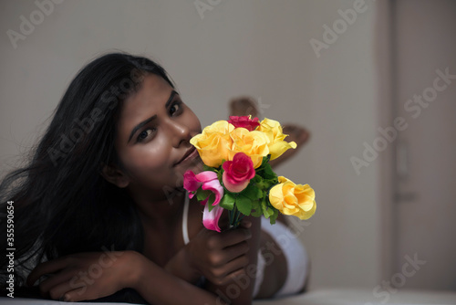 Close up portrait of an young dark skinned Indian Bengali woman in lingerie and vibrant flowers lying on white bed in casual mood in white background. Indian lifestyle and boudoir photography.