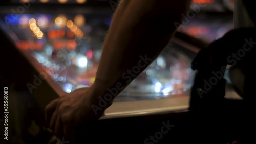 A person playing at a pinball table with the lights flashing out of focus in the background