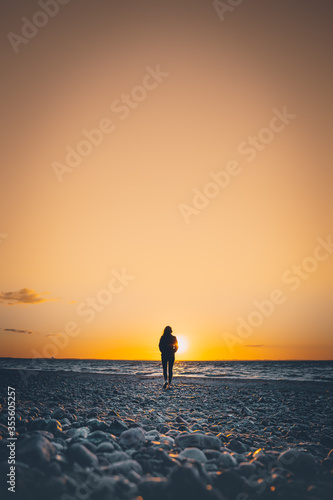 Frau spaziert in Richtung eines wundersch  nen den Sonnenuntergangs an einem steinigen Strand an der Ostsee