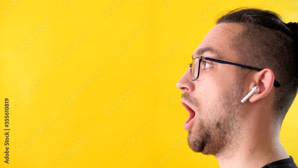 Obraz premium Very surprised man with glasses and headphones looks to the left side of the screen. Profile portrait of an attractive adult unshaven man on a yellow background. Emotional guy opened his mouth