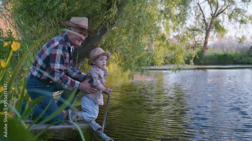 fishermen, caring loving grandfather teaches his little cute grandson to fish with fishing rods in river standing on pier background of reeds and trees fishermen, caring loving grandfather teaches his little cute grandson to fish with fishing rods in river standing on pier background of reeds and trees