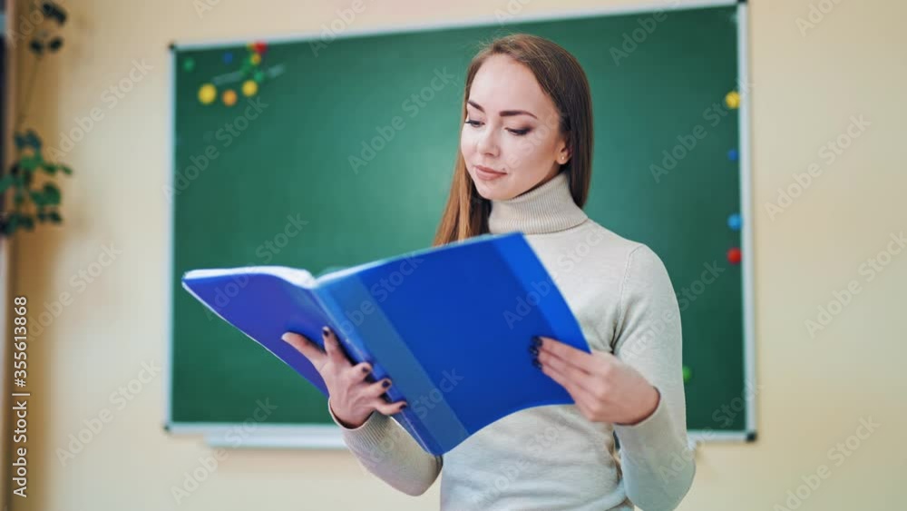 Beautiful teacher with a school register. Portrait of a young female educator looking into the folder on the blackboard background in the classroom. Beautiful teacher with a school register. Portrait of a young female educator looking into the folder on the blackboard background in the classroom.