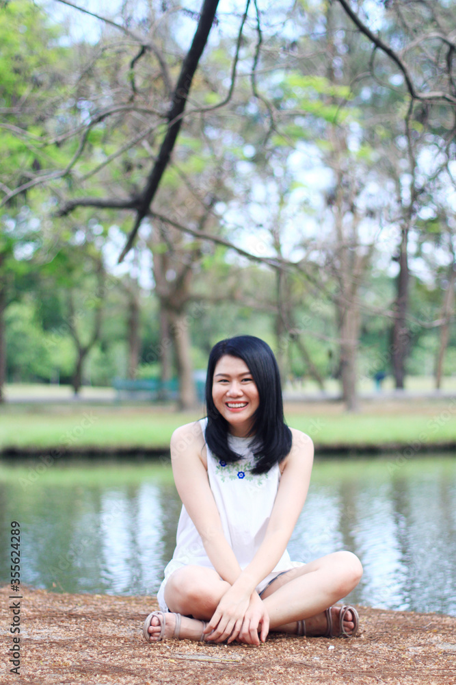 Beautiful asian woman in white dress relax and smiling and sitting on dry field near lake in natural park. Young Thai girl enjoy on holiday with sunlight in garden