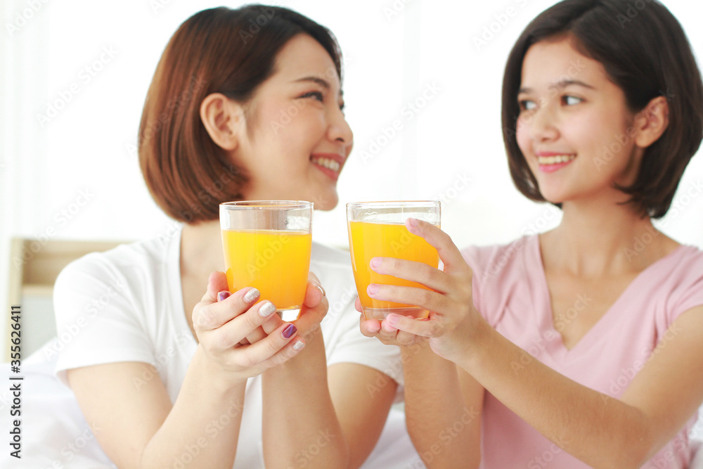 Asian young sisters lovely couple on white bed and smiling with hand holding Orange juice glass in bedroom. Holiday and healthy concept. Closed up on Orange juice glass. Asian young sisters lovely couple on white bed and smiling with hand holding Orange juice glass in bedroom. Holiday and healthy concept. Closed up on Orange juice glass.