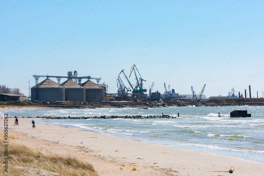 Large grain elevator silos and cranes in port. Sandy beach with people on bikes. (Port of Liepaja, Latvia)