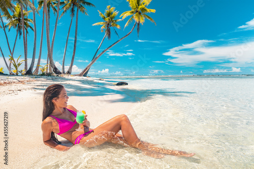 Beach vacation Asian bikini woman enjoying perfect summer holidays drinking blue hawaiian drink lying down sun tanning. Tahiti cruise island relaxing sipping cocktail on French Polynesia travel.