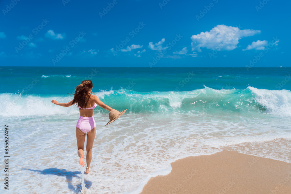 A beautiful girl in a bathing suit with a straw hat in her hand runs towards the ocean. Azure wave rolls on the sandy Paradise beach.
