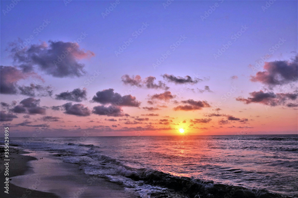 Fantastic sunrise in the Caribbean. The sun has just risen above the horizon. Clouds and sky are painted in incredible scarlet, pink, orange, purple, violet, blue tones. Waves roll to the beach. Fantastic sunrise in the Caribbean. The sun has just risen above the horizon. Clouds and sky are painted in incredible scarlet, pink, orange, purple, violet, blue tones. Waves roll to the beach.