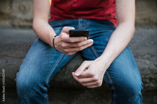 Young man using the smartphone sitting on the bench in the city -Millennial having fun with his friends on social networks - Typing on the touch screen on device for browse the internet